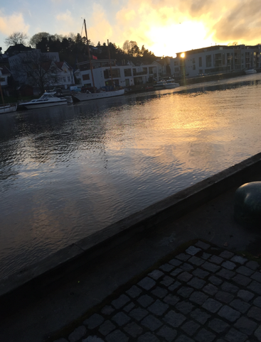 Goldenes Sonnenlicht spiegelt sich auf dem ruhigen Flusswasser. Entlang des Ufers liegen Boote vor Anker. Im Hintergrund reihen sich Häuser und Bäume aneinander und schaffen eine friedliche, malerische Abendstimmung.