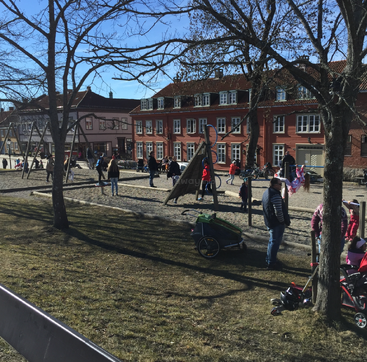 Kinder und Erwachsene genießen einen sonnigen Tag auf einem Spielplatz mit Schaukeln und Klettergerüsten, umgeben von bunten Gebäuden und Bäumen mit kahlen Ästen – es ist Frühling.