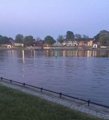 Ein ruhiger Fluss spiegelt die Abendlichter der hübschen Häuser und Bäume am gegenüberliegenden Ufer wider, während eine grüne Boje friedlich im Wasser treibt.