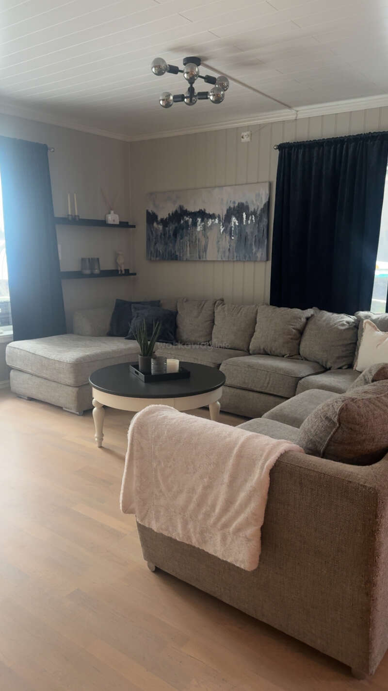 Ein gemütliches Wohnzimmer mit einem großen grauen Ecksofa, einem weißen runden Couchtisch, einem modernen Kronleuchter, abstrakten Kunstwerken, schwarzen Vorhängen und hellem Holzboden. Stilvolle Einrichtung.