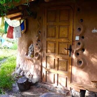 Eine Holztür in einem rustikalen Lehm- oder Steinhaus, verziert mit Gebetsfahnen, kleinen Skulpturen, künstlerischen Wandlöchern, umgeben von Grün und natürlichem Licht.
