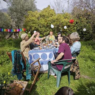 The image depicts a joyful gathering of people and a dog around a table in a lush, grassy outdoor setting, surrounded by trees and festive decorations.