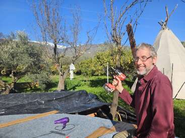 Un homme souriant en plein air tient deux perceuses électriques. Il est entouré d'arbres et d'herbe verte, avec une tente tipi et des montagnes visibles sous un ciel bleu clair.