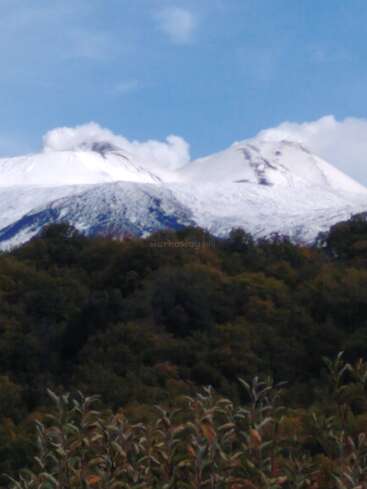 A beautiful landscape features two snow-covered mountains with smoke rising, dense green forest below, and bright blue sky above, creating a serene and majestic scene.