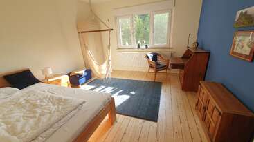 This cozy bedroom features a wooden bed, hammock chair, desk, blue accent wall, wooden furniture, large window, soft lighting, blue rug, and minimalistic, inviting decor.