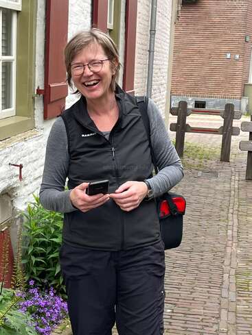 A smiling woman stands on a cobblestone street, holding a phone. She wears glasses, a black vest, and outdoor clothes, appearing cheerful in a charming village setting.