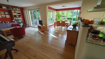 This image shows a spacious living and dining area with large windows, wooden floors, red accents, bookshelves, and a kitchen. Natural light fills the room.