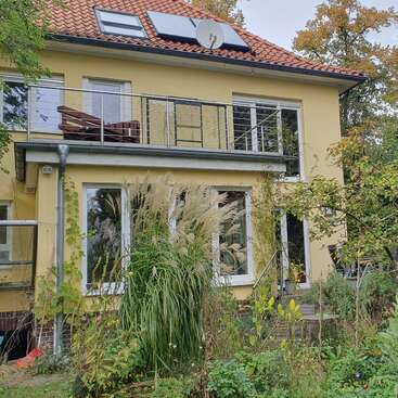 A modern yellow house with a red-tiled roof, solar panels, and a balcony. The lush, green garden in front features ornamental grasses and various plants.