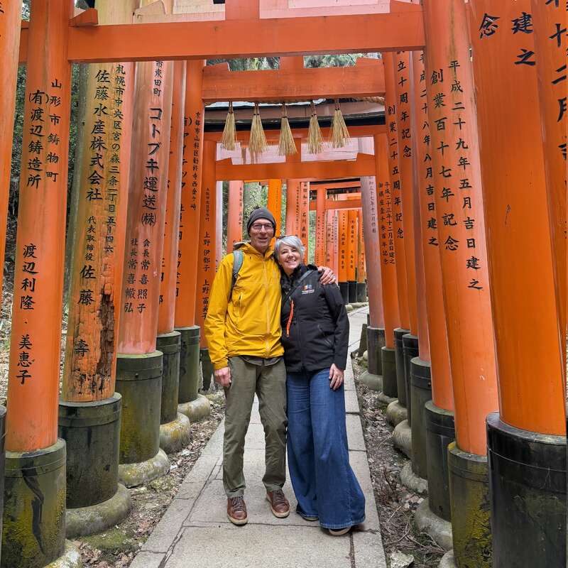Un couple souriant pose ensemble devant des torii d'un orange vif ornés d'inscriptions japonaises, sur l'allée d'un célèbre sanctuaire, dans une atmosphère paisible et imprégnée de culture.