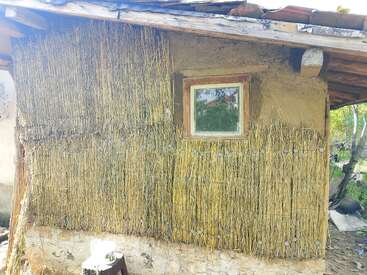 Cette image montre, en extérieur, le mur d'une maison rustique fait de roseaux et de boue, avec une petite fenêtre à cadre en bois, sous un toit en pente en bois.