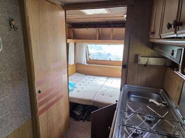 Esta imagen muestra el acogedor interior de una autocaravana, con una cama doble junto a una ventana, una pequeña zona de cocina con fregadero y armarios de madera.