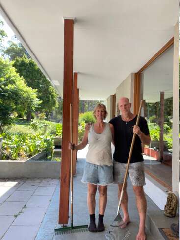 A smiling couple stands on a patio, covered in dirt, holding rakes. They appear to have finished gardening or yard work. Lush green garden surrounds them.