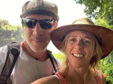A smiling couple is taking a selfie outdoors, surrounded by greenery. Both wear hats; the man has sunglasses and a backpack, suggesting they are on an adventure.