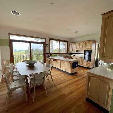 This image shows a spacious kitchen and dining area with wooden floors, light wood cabinets, large windows, a dining table, six chairs, and scenic outdoor views.