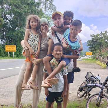 Um grupo alegre de adultos e crianças posam juntos ao ar livre perto de uma estrada, alguns sendo carregados. Há bicicletas por perto, e árvores e montanhas preenchem o fundo.