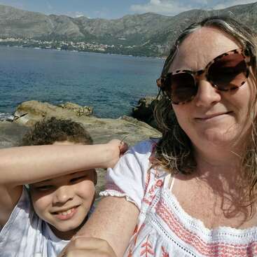 Eine Frau und ein Kind, die beide für ein Selfie am felsigen Meeresufer lächeln. Berge und blaues Wasser bilden unter strahlend blauem Himmel eine wunderschöne Kulisse.