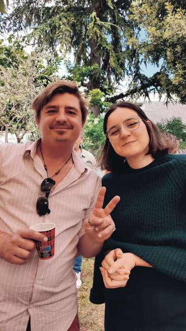 Two people are posing outdoors, smiling at the camera. One holds a cup, the other flashes a peace sign; both seem happy and relaxed among trees.