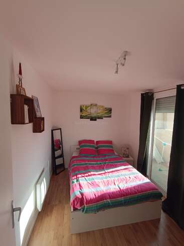 A cozy bedroom with a double bed, colorful striped bedding, wall art above the bed, wooden floors, bookshelf, mirror, window with curtains, and balcony access.