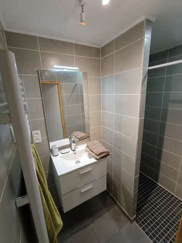 This modern bathroom features gray tile walls, a white vanity with drawers, stacked towels, mirror, overhead light, walk-in shower, and a green towel on the radiator.