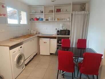 This cozy kitchen has open shelves, a washing machine, small fridge, microwave, coffee maker, and a dining table with four red-covered chairs, and tiled flooring.