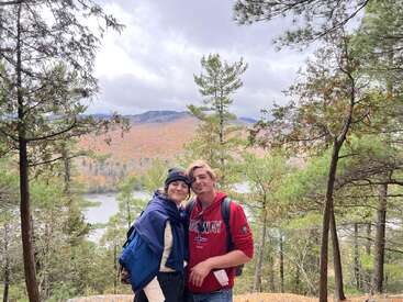 A smiling couple stands together outdoors, surrounded by trees and overlooking a scenic lake with colorful autumn foliage, mountains, and a cloudy sky in the background.