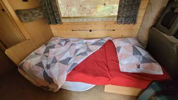 Das Bild zeigt ein gemütliches kleines Bett in einem holzgetäfelten Zimmer mit einer Bettdecke mit geometrischem Muster, einem roten Laken, Kissen, einem Fenster darüber und halb zugezogenen Vorhängen.