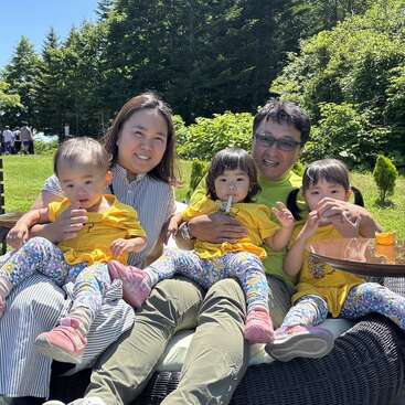 A family of five sits outdoors on a sunny day. The parents and three young girls, all dressed similarly, smile and enjoy snacks together.