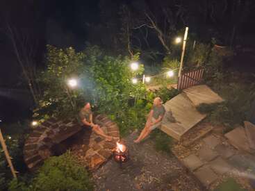 Duas pessoas estão sentadas ao redor de uma pequena fogueira num quintal aconchegante e bem iluminado à noite. Luzes de corda e uma vegetação exuberante criam um ambiente acolhedor e relaxante.