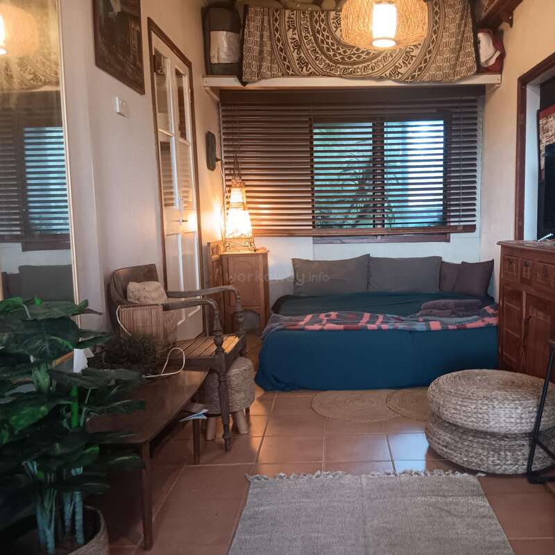 Quarto aconchegante em estilo boêmio, com cama azul, pufes de tecido, móveis de madeira, espelho grande, plantas de interior, iluminação aconchegante, tapeçaria decorativa e persianas. Ambiente acolhedor.