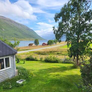 L'image représente un paysage rural serein avec une petite maison, de la verdure luxuriante et un plan d'eau tranquille, sur fond de montagnes pittoresques.