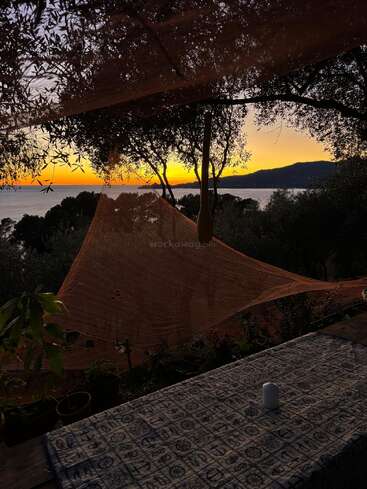 A beautiful sunset over the sea, viewed through trees and netting. Foreground features a table with a patterned cloth and a candle, creating a serene atmosphere.
