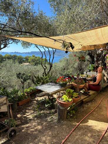 A woman enjoys an outdoor meal under a shaded canopy, surrounded by lush plants, overlooking a beautiful blue sea and distant mountains on a sunny day.