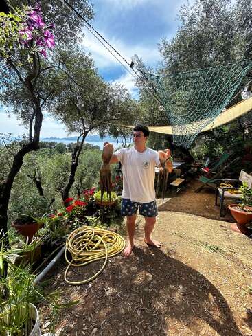 A man in sunglasses stands barefoot outdoors, holding two octopuses. Surrounded by trees, flowers, and garden pots, he’s on a sunny patio with coiled hose nearby.