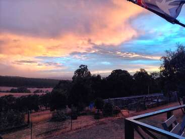 A beautiful sunset paints the sky in vivid pink, orange, and blue hues above a rural landscape with trees, fencing, and a flag in the corner.