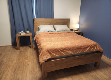 A cozy bedroom features a wooden bed with orange bedding, two side tables, a lamp, a plant, books, blue curtains, and light-colored wooden flooring.
