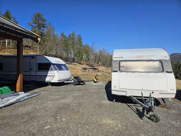 Two caravans are parked on a gravel area next to a wooden structure. Bright blue sky, scattered trees, and hills form the scenic outdoor background.