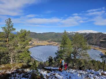 Two hikers overlook a scenic, partially frozen lake surrounded by pine trees. Snow dusts the ground, mountains and a bright blue sky fill the background. Peaceful.