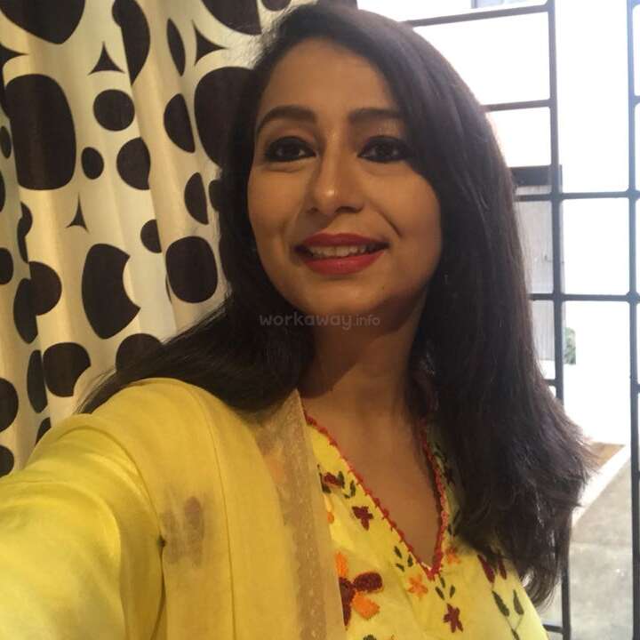 A woman in a yellow floral outfit smiles while taking a selfie indoors. Black-and-white polka dot curtains and window bars are visible in the background.