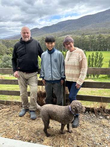 The image depicts a family of three standing in front of a wooden fence, with a dog in the foreground, set against a picturesque mountainous backdrop.
