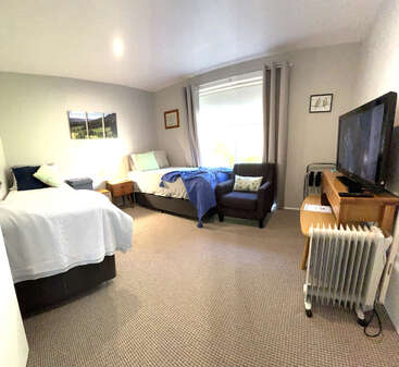 A cozy bedroom featuring two beds, a window with curtains, a TV on a wooden stand, an armchair, artwork, a heater, and neutral decor.