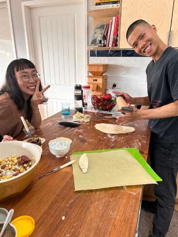 Duas pessoas preparando comida alegremente em uma cozinha, moldando a massa em uma mesa enfarinhada. Ingredientes, utensílios e livros os cercam; uma pessoa faz um sinal de paz.