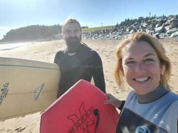 A man and woman in wetsuits smile on a sunny beach, holding surfboards. The ocean, rocks, and trees are visible in the background, creating a fun atmosphere.