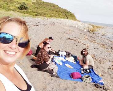 A group of four friends enjoys a beach picnic, sitting on a blue blanket with snacks. One person smiles for a selfie, capturing a sunny, relaxed scene.