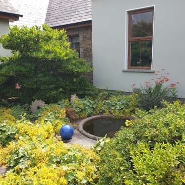 A small garden pond surrounded by lush greenery and yellow flowers, featuring a striking blue sphere ornament, beside a house with stone and plaster walls.