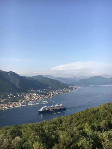 L'image représente une scène côtière sereine, avec un grand navire de croisière naviguant sur un plan d'eau tranquille, entouré d'une végétation luxuriante et de montagnes majestueuses en arrière-plan.
