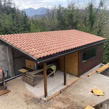 Un petit bâtiment au toit de tuiles rouges est entouré d'arbres. La structure semble en cours de construction, avec des planches de bois, des outils et des matériaux à proximité. Des montagnes en arrière-plan.