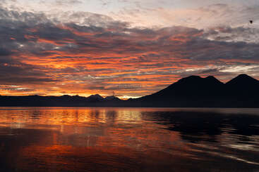 A vibrant sunset illuminates the sky with fiery orange and crimson hues, reflecting beautifully on calm water, while silhouetted mountains create a stunning, tranquil landscape.