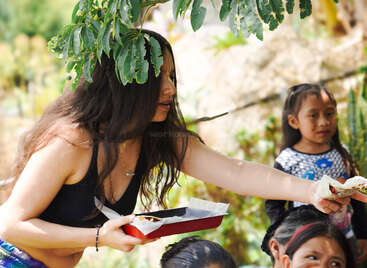 A woman with long hair is serving food outdoors under a tree. Several children are gathered around, and the atmosphere appears warm and friendly.
