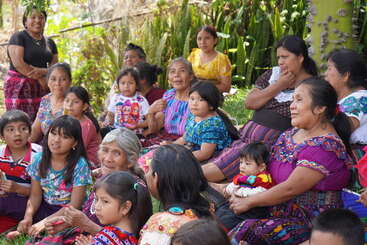A group of women and children, dressed in colorful traditional clothing, sit together outdoors, smiling and conversing, surrounded by lush greenery and natural sunlight.