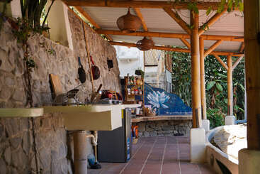 This outdoor kitchen features stone walls, bamboo beams, hanging pots, a small fridge, and natural light. Plants and a large lotus mural create a peaceful atmosphere.